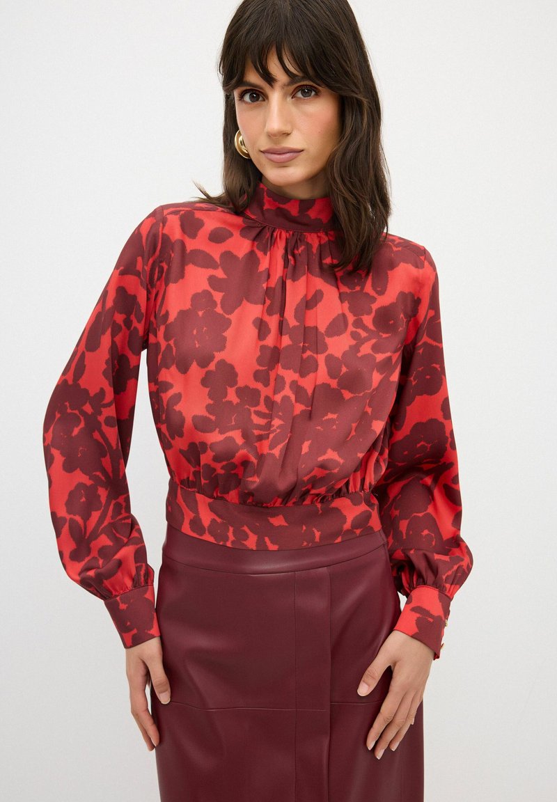 Rode blouse met bloemenpatroon, lange mouwen, hoge kraag en plooidetails, gecombineerd met een bordeauxlederen kokerrok.