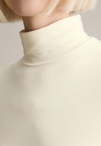 Cremfarbener Rollkragenpullover aus glattem, dehnbarem Material. Nahaufnahme zeigt das Design des Kragens und die feine Textur, die die taillierte Silhouette betont.