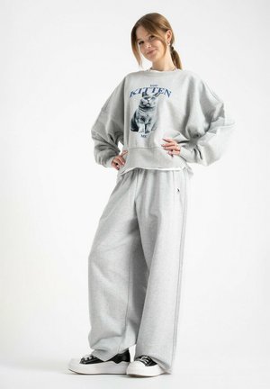 Młoda kobieta z kucykiem, ubrana w luźną szarą bluzę z nadrukiem kota oraz dopasowane szerokie spodnie, stoi z rękami na biodrach i uśmiecha się.