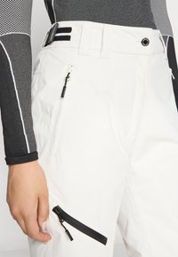 Hvite skibukser med teksturert overflate, med en svart glidelåslomme, svarte beltesløyfer, og en glatt midje med sømdetaljer.