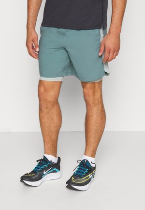 Mænds atletiske shorts i teal med en lys grå talje, parret med sorte og blå løbesko med en hvid sål og hvide sokker.