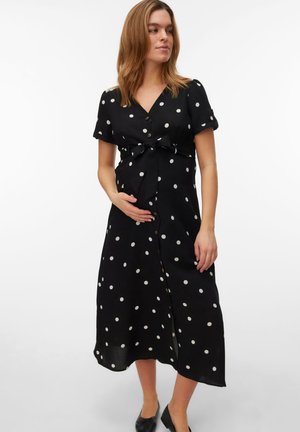 Gravid kvinde i sort midi-kjole med hvide prikker, korte ærmer og knapper, der blidt holder sin mave, stående mod en hvid baggrund.