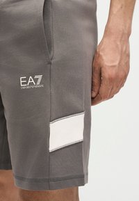 Κοντινό πλάνο του χεριού ενός ατόμου δίπλα σε γκρι σορτς με λογότυπο Emporio Armani EA7 και λευκή οριζόντια ρίγα κοντά στο τελείωμα.