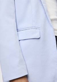 Světle modrý blazer s hladkým materiálem, jednou přední kapsou na levé straně a upravenými rukávy pro strukturovaný siluetu.