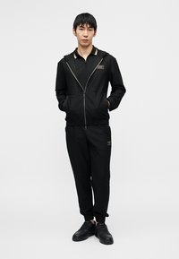 Μαύρο φούτερ με φερμουάρ από υφή υλικό με χρυσές λεπτομέρειες, συνδυασμένο με ταιριαστά παντελόνια φόρμας, και τα δύο με στενή γραμμή και ελαστικά μανσέτες.