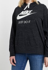 Sudadera negra con tejido texturizado, bolsillo frontal tipo canguro, logo blanco de Nike y texto "JUST DO IT", con capucha ajustable con cordón.