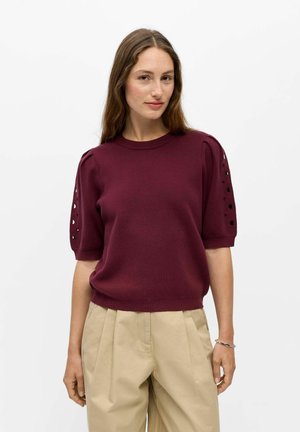 Femme aux longs cheveux bruns portant un haut en maille bordeaux à manches courtes avec des détails ajourés et un pantalon plissé beige sur fond blanc.