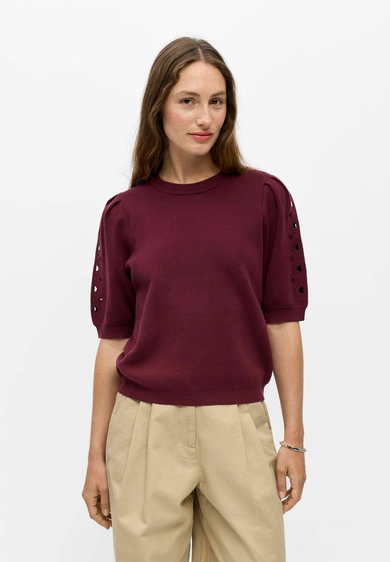 Femme aux longs cheveux bruns portant un haut en maille bordeaux à manches courtes avec des détails ajourés et un pantalon plissé beige sur fond blanc.