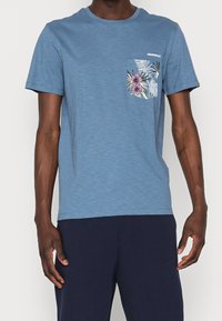 Modré krátké tričko s krátkým rukávem vyrobené z kombinace bavlny, s kapsou s květinovým vzorem v purpurové a zelené barvě.