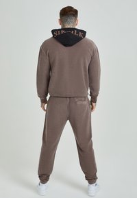 Brun fleece-träningsoverall med svart huva. Huvan har en logotyp på kragen. Slim-fit byxor med en ficka på ryggen. Vita sneakers.