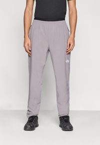 Γκρι outdoor παντελόνι με ελαστική μέση, με τσέπες στο πλάι και λογότυπο στο αριστερό μηρό. Φορεμένο με μαύρα αθλητικά παπούτσια.