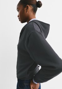 Felpa nera con cappuccio dal taglio morbido. Tessuto morbido, polsini a coste e chiusura con zip. Indossata sopra una camicia a righe con colletto.