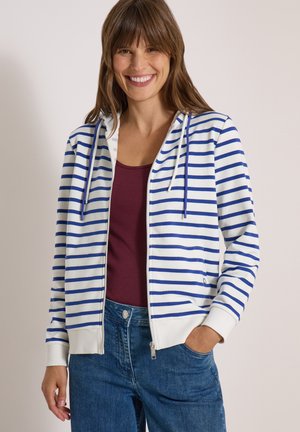 Femme souriante portant un sweat à capuche zippé blanc et bleu à rayures horizontales, un haut bordeaux et un jean bleu avec une main dans la poche.