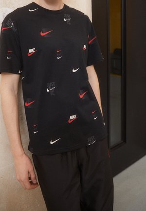 Osoba nosząca czarną koszulkę Nike z czerwonymi i białymi logo typu swoosh oraz napisem „Just Do It”, w połączeniu z czarnymi spodniami.
