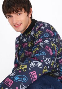 Giacca bomber con sfondo blu navy e una stampa grafica di controller e console di gioco in giallo, rosa e blu. Collo nero.