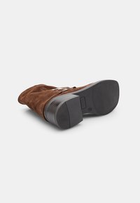 Stivale alla caviglia in suede marrone con tacco a blocco, punta curva e suola nera liscia. Caratterizzato da cuciture minime e un sottile accento con logo.