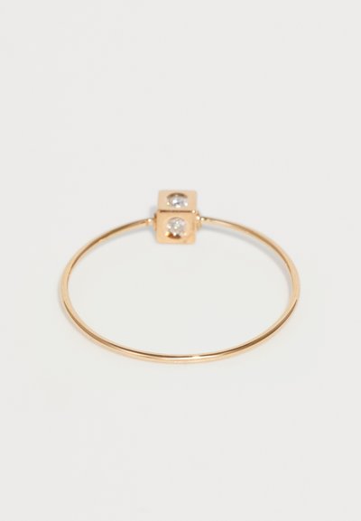 Anillo de metal en tono dorado con una banda delgada. Presenta un acento en forma de cubo con dos piedras preciosas transparentes en la parte superior. Diseño simple y geométrico.