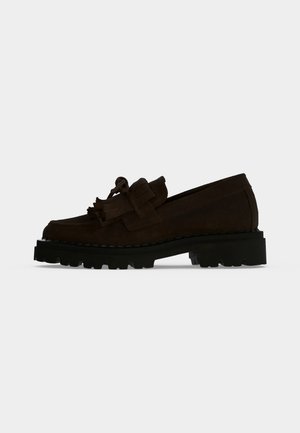 Mocassin en daim marron foncé avec semelle crantée noire épaisse et détail pompon sur le dessus, présenté de profil sur un fond clair uni.