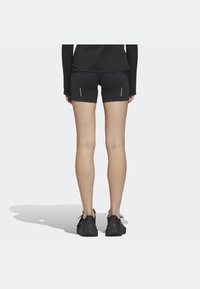 Osoba stojąca tyłem, ubrana w czarne spodenki sportowe z odblaskowymi paskami oraz czarne buty do biegania na jasnym tle.