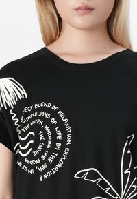 Juodas medvilnės marškinėliai su baltais grafikos elementais: delno lapai ir spiralinis tekstas. Apvalus kaklas ir trumpi rankovės.