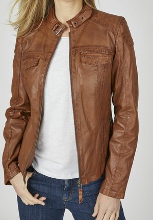 Vrouw draagt een bruine leren jas over een wit overhemd en blauwe spijkerbroek, met de jas gedeeltelijk open en een hand in haar zak.