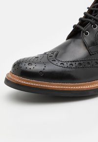 Zapato brogue de cuero negro con detalles perforados, punta redonda y un diseño clásico de cordones. Acentos en la media suela de cuero marrón.