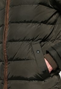Oliven grøn pufferjakke med brun lynlås og kant. Har quiltede sektioner, en lomme med trykknaplukning og ribbede manchetter.