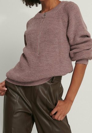 Person trägt einen mauvefarbenen Strickpullover mit einem Reißverschluss vorne und dunkelbraune, hoch taillierte Lederhosen und steht mit einer Hand in der Tasche.
