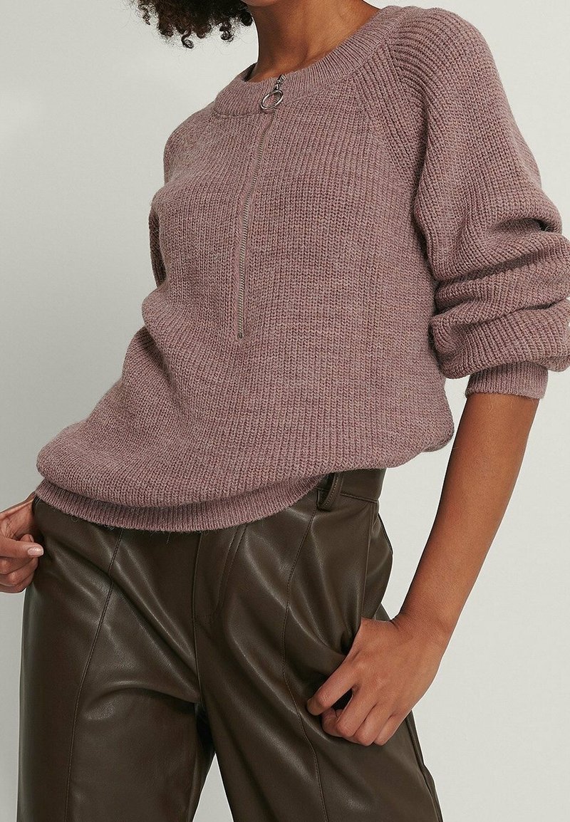 Personne portant un pull en maille mauve avec une fermeture éclair à l'avant et un pantalon en cuir marron foncé taille haute, debout avec une main dans la poche.