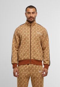Brown patterned zip-up jacket with high collar, ribbed cuffs, and waistband, featuring a contrasting navy side stripe and logo detail.