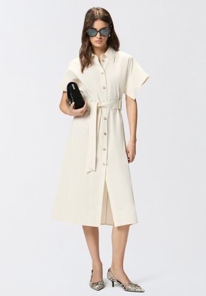 Femme portant une robe midi beige boutonnée avec ceinture à la taille, lunettes de soleil noires, tenant une pochette noire et portant des talons à motif peau de serpent.