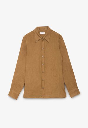 Femme aux cheveux foncés attachés, portant une chemise marron boutonnée et un collier chaîne en or épais sur un fond uni.