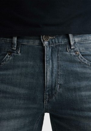 Nahaufnahme einer Person, die dunkelblaue Jeans mit Metallknopf und Nieten trägt, kombiniert mit einem schwarzen Hemd, das leicht in den Hosenbund gesteckt ist.
