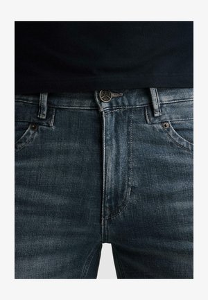 Nahaufnahme einer Person, die dunkelblaue Jeans mit Metallknopf und Nieten trägt, kombiniert mit einem schwarzen Hemd, das leicht in den Hosenbund gesteckt ist.