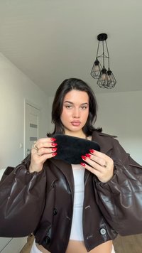 Žena s tmavými vlasy a červenými nehty držící černou masku na obličej, oblečená v hnědé kožené bundě a bílem topu, stojící uvnitř pod černým závěsným světlem.