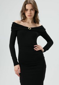 Schwarzes schulterfreies Kleid aus elastischem Stoff mit gerafftem Design, langen Ärmeln und einer goldenen ovalen Brosche als Akzent.