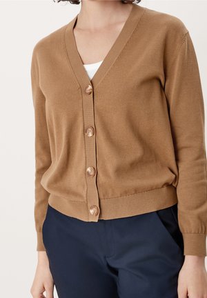 Personne portant un cardigan marron boutonné à col en V sur un haut blanc, associé à un pantalon bleu marine, montré du cou aux hanches.