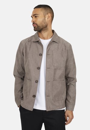 Mężczyzna z brodą w zapiętej taupe zamszowej kurtce, białej koszuli i czarnych spodniach, patrzący w lewo, z rękami w kieszeniach.