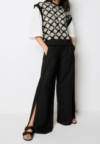 Un pull surdimensionné à motifs noir et blanc sur un haut à manches bouffantes blanc, associé à un pantalon large noir avec des fentes sur les côtés et des sandales noires.