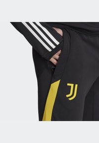 Schwarze Sporthose mit gelbem Streifen und Logo-Details; verfügt über ein weißes Drei-Streifen-Design auf dem Ärmel; glatter Stofftextur.