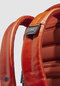 Correa de mochila de cuero marrón con un forro de tela texturizada, que incluye una etiqueta metálica marcada con "M.P." y acentos en azul contrastante.