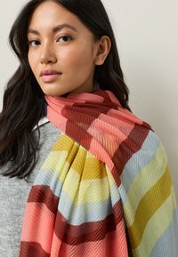 Bufanda de rayas multicolores con textura acanalada, que presenta tonos rojos, rosas, amarillos y azules. Colgada sobre un suéter gris y un cuello.