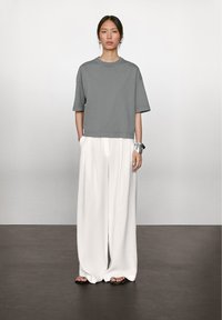 Grå kortærmet sweatshirt parret med løse hvide brede bukser. Modellen bærer et sølvarmbånd og flade sorte sandaler på et mørkt gulv.