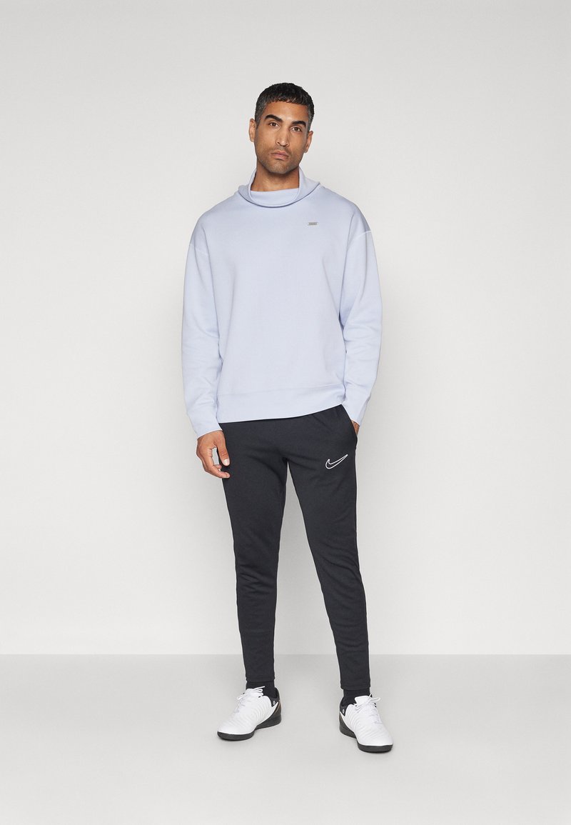 Felpa azzurro chiaro con collo alto e dettaglio logo argento, abbinata a pantaloni della tuta neri con dettagli attillati e logo Nike bianco. Sneakers bianche.