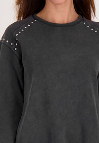 Vrouw in een donkergrijze sweatshirt met zilveren studs langs de schoudernaden en bovenste mouwen, weergegeven van hals tot midden-torso.