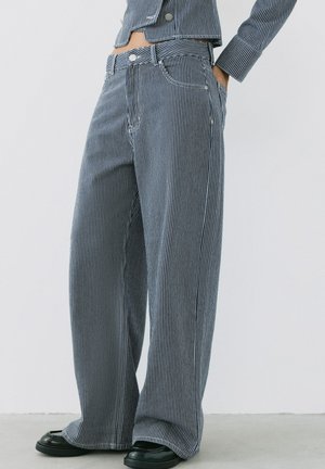 Personne portant un pantalon ample rayé verticalement bleu et blanc, mains dans les poches, et une veste assortie, debout sur un sol gris.