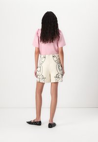 Haut rose à manches courtes avec épaules bouffantes ; short crème avec motifs de gribouillis noirs ; chaussures plates noires avec quincaillerie décorative.