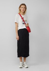 T-shirt blanc avec un graphique de palmier et le texte "IBIZA", jupe midi plissée noire, sac bandoulière rouge et baskets de couleur claire.