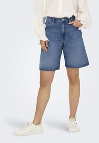 Jeans kratke hlače v srednji modri barvi, z ohlapnim krojem in surovim robom. Ujemajo se z belo bluzo dolgega rokava in svetlimi supergami.