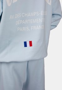 Hellblaues Sportoutfit mit weißen Textdetails. Verfügt über eine kleine gestickte französische Flagge und ein Emblem an der unteren Seite.